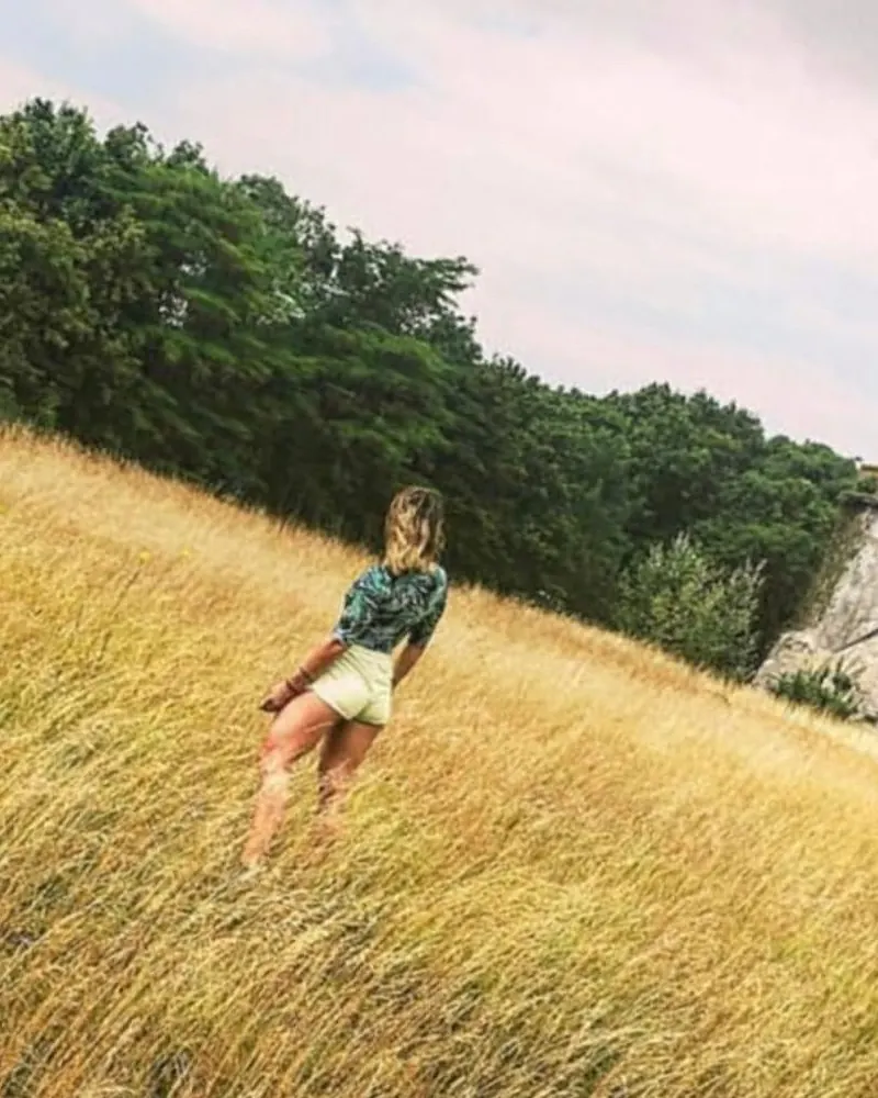 Femme vue de dos marchant dans des herbes hautes dorées, au bord d’un bois, par un jour d’été